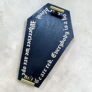 May include: A navy blue coffin-shaped serving tray with gold-coloured handles. The tray has white gothic lettering around the edges that reads "Wherever we are open, we are red. Everybody is a blood."