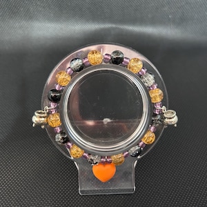 May include: A beaded bracelet with orange, black, silver, and gold beads. The bracelet has a small orange heart charm and silver cauldron charms. The bracelet is displayed on a clear plastic stand.