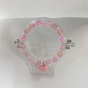 May include: A pink beaded bracelet with a heart charm. The bracelet features pink and clear beads, with silver-colored bow accents. The heart charm is a solid pink color. The bracelet is displayed on a clear plastic stand.