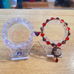 May include: Two beaded bracelets displayed on clear stands. One bracelet is purple and white, with a heart and wing charm. The other is red and black, with a crescent moon charm. Both bracelets are connected by a heart.