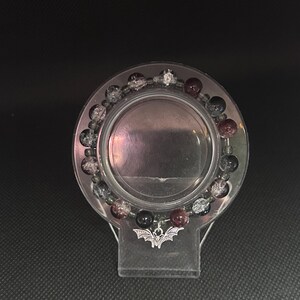 May include: A beaded bracelet with a bat charm. The bracelet features a mix of black, clear, and burgundy-colored beads. The charm is a silver-colored bat. The bracelet is displayed on a clear, circular stand.