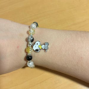 Może przedstawiać: Bransoletka z koralik&oacute;w z zawieszką Snoopy. Bransoletka ma przezroczyste, srebrne, ż&oacute;łte i czarne koraliki. Zawieszka Snoopy jest biała, czarna i ż&oacute;łta. Bransoletka jest na nadgarstku.