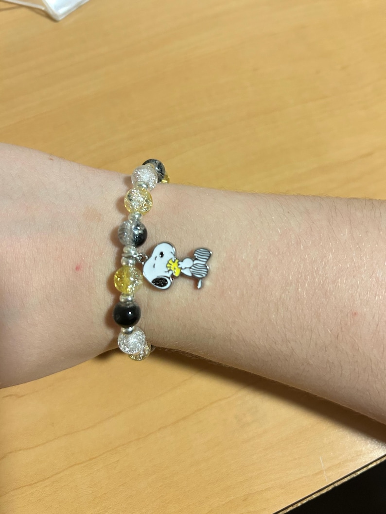 Może przedstawiać: Bransoletka z koralik&oacute;w z zawieszką Snoopy. Bransoletka ma przezroczyste, srebrne, ż&oacute;łte i czarne koraliki. Zawieszka Snoopy jest biała, czarna i ż&oacute;łta. Bransoletka jest na nadgarstku.