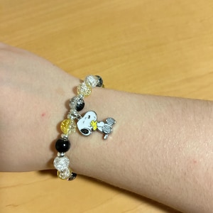 Może przedstawiać: Bransoletka z czarnych, ż&oacute;łtych i przezroczystych koralik&oacute;w. Posiada zawieszkę Snoopy, przedstawiającą postać z kresk&oacute;wki. Bransoletka jest noszona na nadgarstku.
