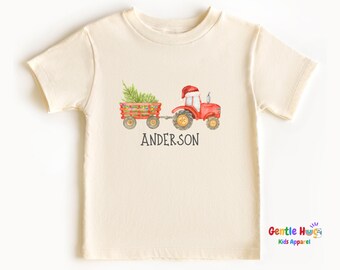 May include: Cream-colored t-shirt featuring a watercolor illustration of a red tractor pulling a wagon filled with a Christmas tree and lights. The tractor has a Santa hat. The name "ANDERSON" is printed below the tractor.