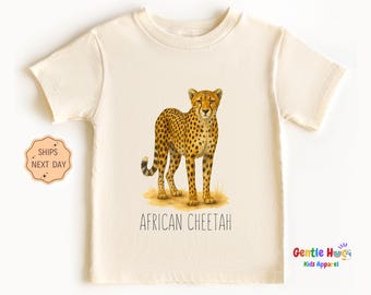 May include: Cream-colored t-shirt featuring a detailed illustration of an African cheetah. The cheetah is depicted with its distinctive spotted coat and is standing on a patch of grass. The text "AFRICAN CHEETAH" is printed below the image.