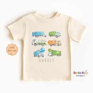 May include: Cream-colored t-shirt featuring a watercolor illustration of six different garbage trucks in various colors. The name "HARVEY" is printed in black below the trucks. The shirt is from Gentle Hugs Kids Apparel.
