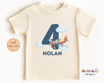 May include: Cream-colored toddler t-shirt featuring a blue number 4, a watercolor airplane, and the name "NOLAN" in blue. The airplane is orange and blue with white clouds. The shirt is short-sleeved.