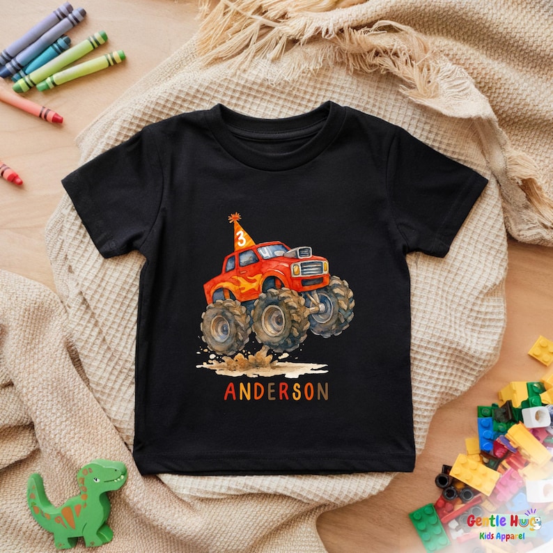 以下が含まれることがあります： カラフルなモンスタートラックのデザインが施された黒い半袖Tシャツ。トラックは赤色で、炎と大きなタイヤがあり、3の数字が入ったバースデーハットをかぶっています。「ANDERSON」という名前が下にプリントされています。子供服。
