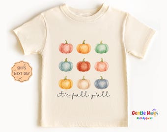 May include: Cream-colored short-sleeved t-shirt featuring a watercolor design of nine pumpkins in various colors. The phrase "it's fall y'all" is printed below the pumpkins. The shirt is from Gentle Hugs Kids Apparel.