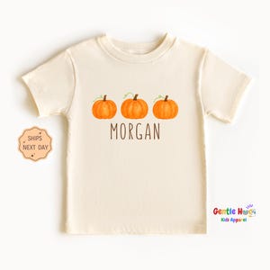 May include: Cream-colored t-shirt featuring three orange pumpkins with green vines above the word "MORGAN" in brown lettering. The shirt is from Gentle Hugs Kids Apparel.