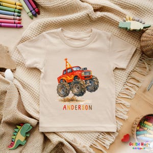 以下が含まれることがあります： ベージュ色のTシャツ。赤いモンスターカーの大きなタイヤ、誕生日の帽子、数字の3のイラストが水彩画で描かれています。「ANDERSON」という名前がカラフルな文字で下に印刷されています。子供向けのTシャツです。