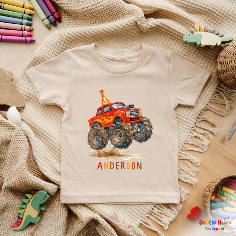 以下が含まれることがあります： ベージュ色のTシャツ。赤いモンスターカーの大きなタイヤ、誕生日の帽子、数字の3のイラストが水彩画で描かれています。「ANDERSON」という名前がカラフルな文字で下に印刷されています。子供向けのTシャツです。