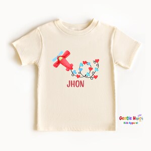 May include: Cream-colored t-shirt featuring a red airplane with blue propellers and a trail of red and blue hearts. The name "JHON" is printed in red below the design. The shirt is from Gentle Hugs Kids Apparel.