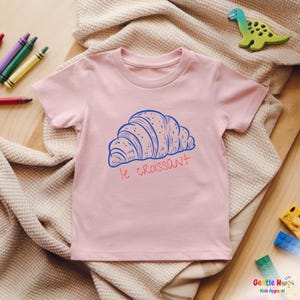 May include: A light pink t-shirt featuring a blue illustration of a croissant with the text "le croissant" in red. The shirt is made of soft material and is suitable for kids. The shirt is laying on a beige blanket.