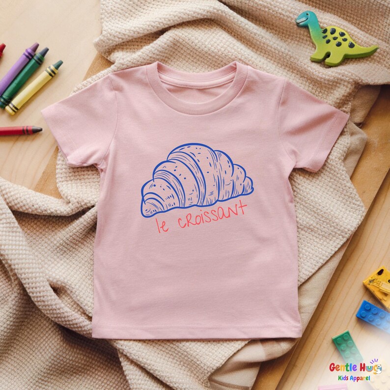 May include: A light pink t-shirt featuring a blue illustration of a croissant with the text "le croissant" in red. The shirt is made of soft material and is suitable for kids. The shirt is laying on a beige blanket.
