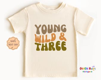 May include: Cream-colored toddler t-shirt with the text "YOUNG WILD & THREE" in brown, orange, and green. The shirt is a short-sleeved crew neck. The shirt is made of a soft material.