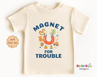 May include: Cream-colored toddler t-shirt with the words "MAGNET FOR TROUBLE" in blue. The design includes a red horseshoe magnet with toy illustrations. A small sticker on the left says "SHIPS NEXT DAY".