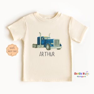 May include: Cream-colored short-sleeved t-shirt featuring a blue watercolor illustration of a semi-truck. The name "ARTHUR" is printed in black below the truck. The shirt is from Gentle Hugs Kids Apparel.