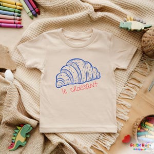 May include: Beige short-sleeved t-shirt featuring a blue illustration of a croissant with the text "le croissant" in red. The shirt is part of a kids apparel collection.
