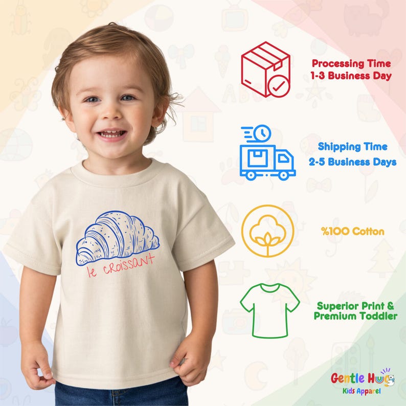 May include: A toddler wearing a beige t-shirt with a blue and red graphic of a croissant and the text "le croissant". The shirt is made of 100% cotton and is part of the Gentle Hug Kids Apparel collection.