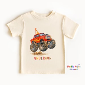 以下が含まれることがあります： クリーム色の幼児用Tシャツ。大きなタイヤと誕生日の帽子をかぶった赤いモンスタートラックの水彩イラストが描かれています。帽子には数字の「3」が描かれています。「ANDERSON」という名前がオレンジ色の文字でトラックの下に印刷されています。