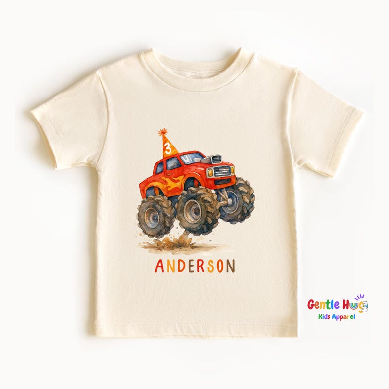 以下が含まれることがあります： クリーム色の幼児用Tシャツ。大きなタイヤと誕生日の帽子をかぶった赤いモンスタートラックの水彩イラストが描かれています。帽子には数字の「3」が描かれています。「ANDERSON」という名前がオレンジ色の文字でトラックの下に印刷されています。