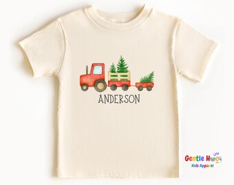 May include: Cream-colored short-sleeved t-shirt featuring a watercolor illustration of a red tractor pulling two wagons filled with Christmas trees. The name "ANDERSON" is printed below the tractor in black.