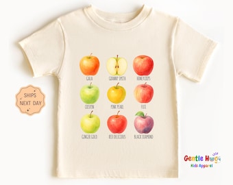 May include: Cream-colored short-sleeved t-shirt featuring a watercolor illustration of nine different apple varieties, each labeled with its name. The shirt also has the text "Gentle Hugs Kids Apparel".