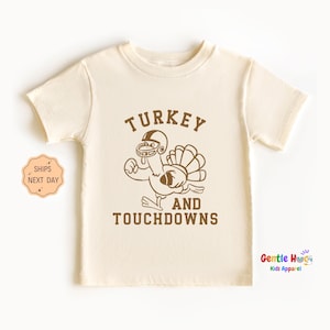 May include: Cream-colored t-shirt featuring a brown cartoon turkey wearing a football helmet, holding a football, and with its tail feathers spread. The text "TURKEY AND TOUCHDOWNS" is above and below the turkey graphic. The shirt is from Gentle Hugs Kids Apparel.