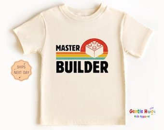 May include: Cream-colored short-sleeved t-shirt with the words "MASTER BUILDER" in black block letters. A red and orange circle with a white and gray building block graphic is above the word "BUILDER". The shirt has a retro design.