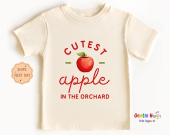 May include: Cream-colored toddler t-shirt with the words "Cutest Apple in the Orchard" in red script and a red apple graphic. The shirt is made of soft material and has short sleeves.
