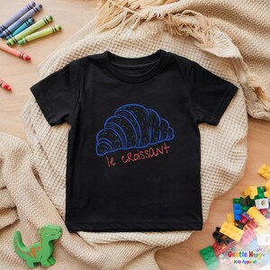 May include: Black t-shirt featuring a blue croissant graphic with the text "le croissant" in red. The shirt is made of a soft material and is designed for kids. The image also includes crayons and toy blocks.