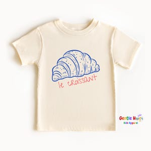 May include: Cream-colored t-shirt featuring a blue line drawing of a croissant. The words "le croissant" are written in red beneath the pastry. The shirt is from Gentle Hugs Kids Apparel.