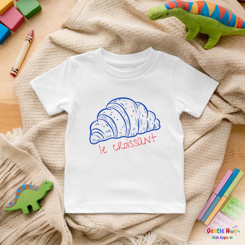 May include: White t-shirt featuring a blue and red illustration of a croissant with the text "le croissant". The shirt is designed for kids and is surrounded by toys.