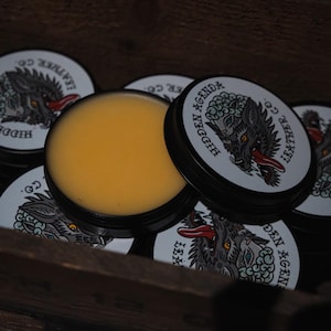 May include: A collection of small, round tins with black lids and white labels. The labels feature a wolf illustration with the text "Hidden Agenda Leather Co." The tins are filled with a yellow balm, and are arranged in a wooden box.
