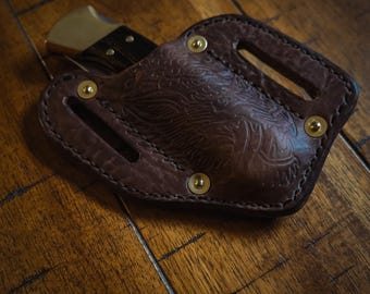 Peut inclure: Un étui à couteau en cuir marron avec un motif gaufré décoratif et des rivets en laiton. L'étui est conçu pour contenir un couteau pliant.