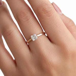 May include: A gold band ring with a square-cut, clear gemstone. The ring is simple and elegant, showcasing the gemstone's brilliance. The ring is shown on a person's finger.