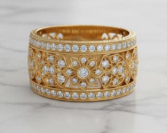 May include: A gold ring with a floral design and embedded diamonds. The ring has a wide band with intricate cut-out patterns and is encrusted with numerous small diamonds, creating a sparkling effect. The ring is set against a marble surface.