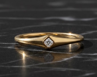 May include: A gold ring with a small, square-cut diamond set in a bezel. The ring has a simple, elegant design with a slightly elongated band. The ring is displayed on a black marble surface.