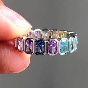 May include: A silver ring featuring a series of colorful, rectangular gemstones. The gemstones are arranged in a row, showcasing a spectrum of colors including pink, purple, blue, and green. The ring is held up against a gray background.
