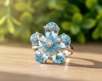 May include: A silver ring featuring a flower design with five light blue gemstone petals. The center of the flower has a small, round, clear gemstone. The ring is set against a blurred green background.