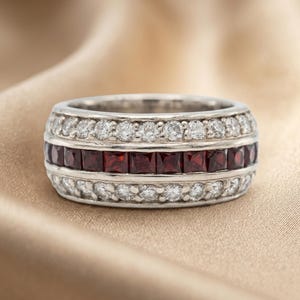 May include: A silver-toned band ring featuring a central row of square-cut deep red gemstones flanked by two rows of small, round, clear white stones. The metal band has a polished finish and a wide, uniform profile across the entire circumference.