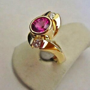 May include: A gold ring featuring a large, oval-cut pink gemstone. Two smaller diamonds flank the main stone. The ring has a unique, flowing design with polished gold surfaces.