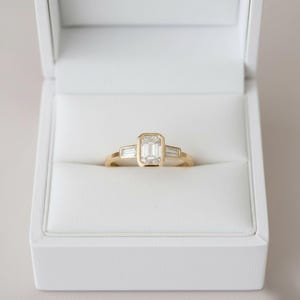May include: A gold ring with an emerald-cut center stone and two baguette-cut side stones, set in a white box. The ring has a simple, elegant design, perfect for an engagement or special occasion.