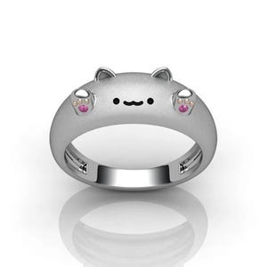 May include: A silver-colored ring with a cat face design. The ring features small ears, a simple black mouth, and pink paw prints on each side. The ring is a cute and whimsical piece of jewelry.