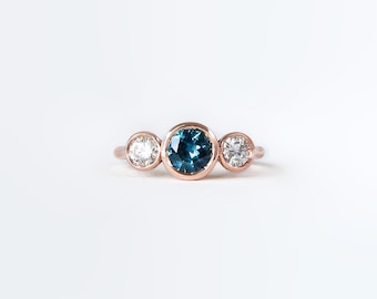 May include: A rose gold ring with three gemstones. The center stone is a round, deep teal color, flanked by two smaller, clear, round gemstones. The ring is set against a white background.