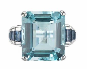 May include: A square-cut, light blue gemstone ring with smaller blue stones on the sides. The ring has a silver band and four silver prongs holding the large gemstone. The ring is a statement piece.