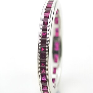 May include: A silver ring with a continuous row of square-cut, deep red gemstones. The ring has a detailed, textured border on either side of the gemstones. The ring is set against a white background, highlighting the vibrant color of the stones.