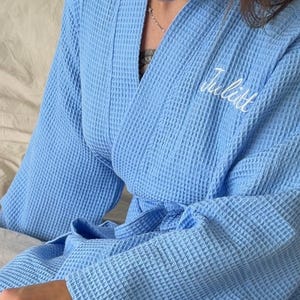 May include: A light blue waffle-knit robe featuring long sleeves, a tie waist, and the name "Julitt" embroidered in white script on the upper left chest. The textured fabric has a grid-like pattern throughout the garment.